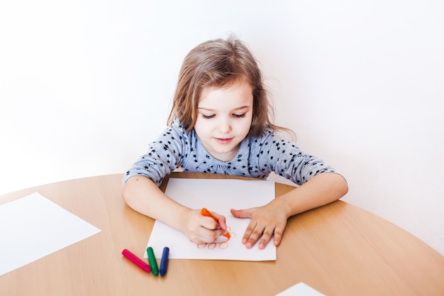 A little girl in a gray dress sits at a table and draws a flower with wax crayons