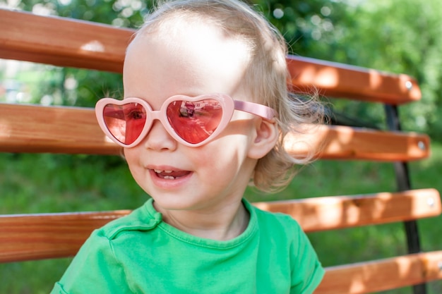 Photo little girl in a green t-shirt and pink heart-shaped sunglasses walks in the park in summer. high quality photo