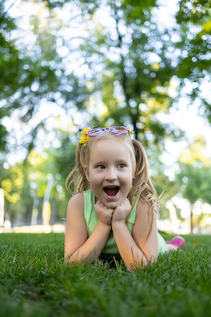 A little girl in a green tracksuit lies on the lawn in the park pressing her hands to her chin and rejoicing