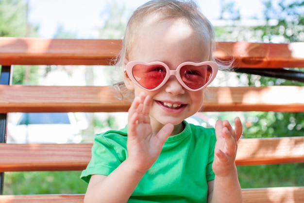 Una bambina con una maglietta verde e occhiali da sole rosa a forma di cuore cammina nel parco in estate