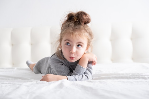 Little girl in grey turtleneck having fun on white bedding.