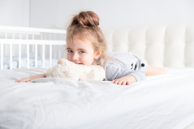 Little girl in grey turtleneck playing with teddy bear on white bedding.