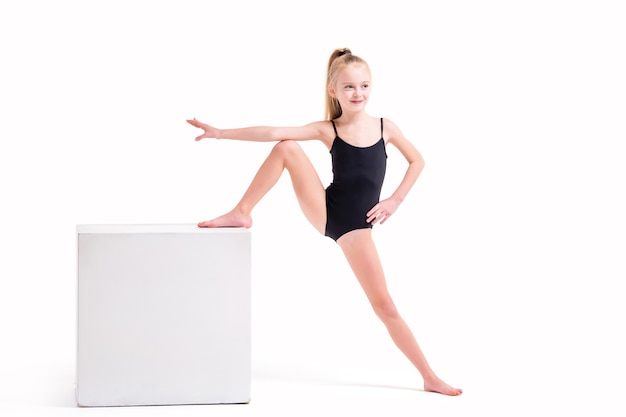 Фото Маленькая девочка гимнастка в черном купальнике стоя одной ногой на белом кубе, изолированном на белом фоне