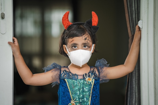 Little girl in halloween costume wearing protective face mask standing indoor