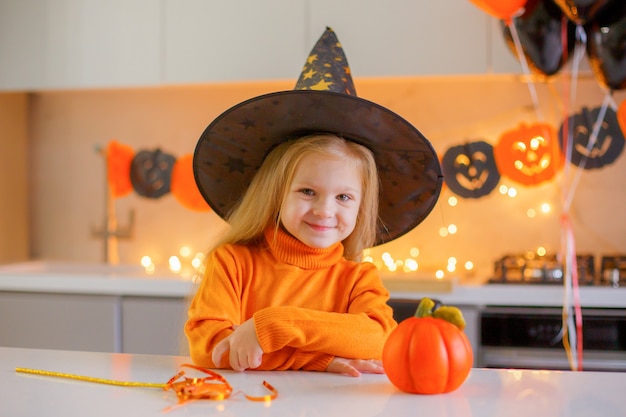 Little girl on Halloween in a witch costume with a pumpkin at home in the kitchen