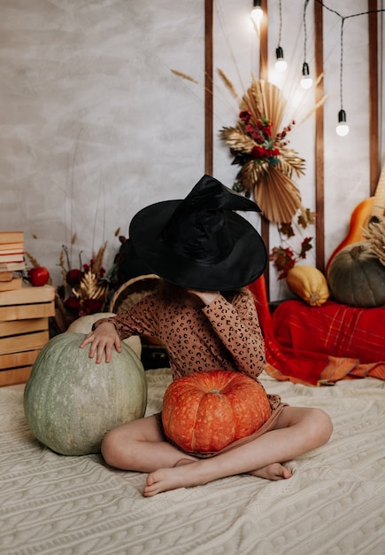 A little girl has covered her face with a witch's hat and is sitting on a knitted blanket with pumpkins