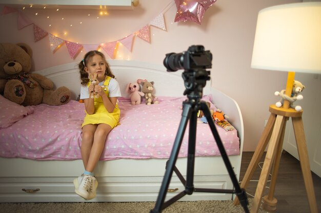 A little girl has her own video blog. he talks about his toys\
on a digital video camera. communicates live stream with his\
followers. shows them sitting in his room at home on the bed.