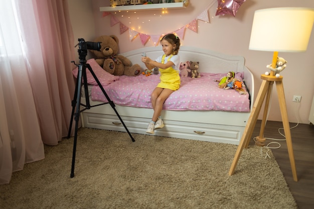 A little girl has her own video blog. he talks about his toys\
on a digital video camera. communicates live stream with his\
followers. shows them sitting in his room at home on the bed.