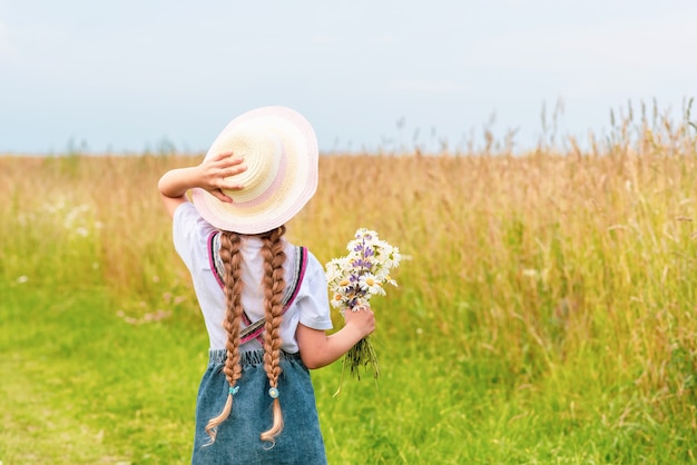 Маленькая девочка в шляпе держит букет ромашек и смотрит в поле