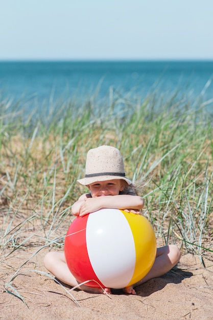 Little girl in hat playing on beach with ball, sunny summer day. Holiday concept