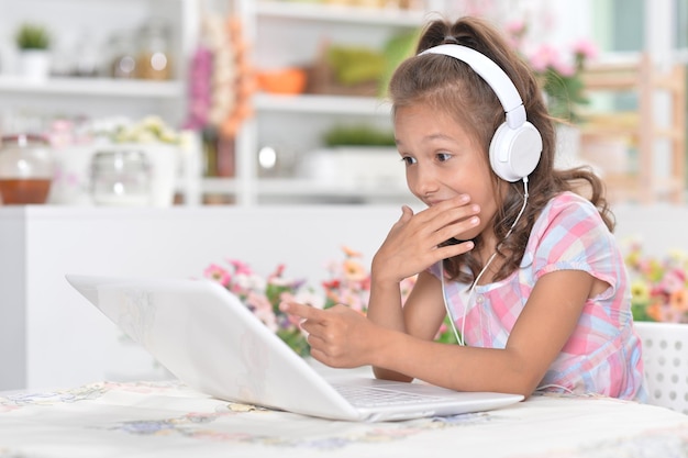 Little girl in headphones sitting at table and using laptop