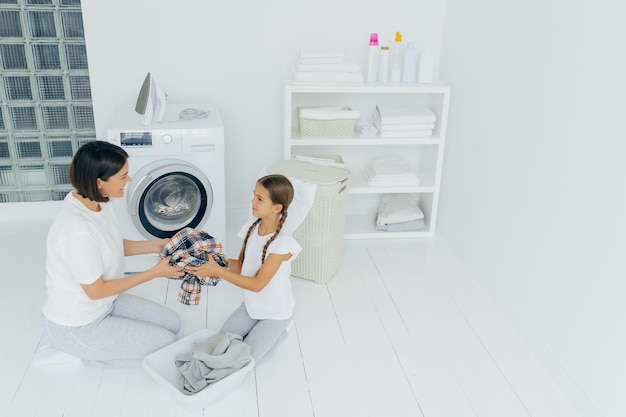 Little girl helper and her mother pose in laundry room near\
washing machine sit on white floor hold dirty shirt do washing at\
home during weekend load clothes family chores and domestic\
work