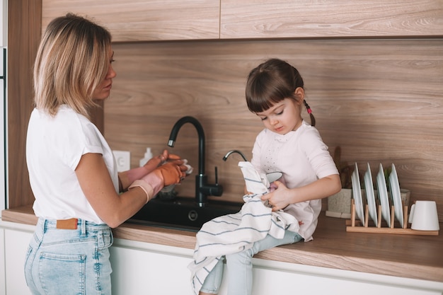 부엌에서 주방용으로 그녀의 어머니를 돕는 어린 소녀. 여자가 설거지를하고 있고 딸이 수건으로 컵을 닦고 있습니다.