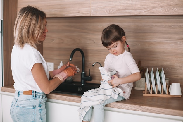 Little girl helping her mother with dishwashing in the kitchen. Woman is washing the dishes, her daughter wiping the cup off with a towel.