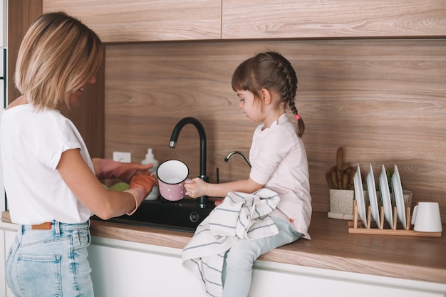 Bambina che aiuta sua madre con il lavaggio dei piatti in cucina. la donna lava i piatti, sua figlia pulisce la tazza con un asciugamano.