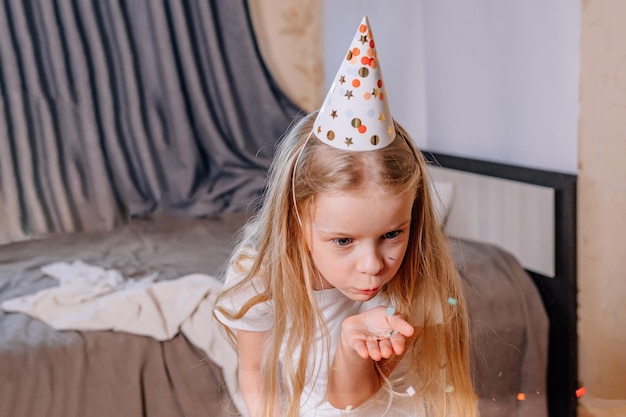 Little girl on her birthday morning inflates multicolored
confetti from palm her hand in festive cap a child celebrates his
birthday at home during illness quarantine isolation holiday
without guests