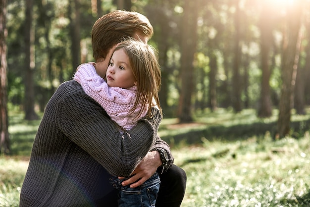 Bambina e suo padre nella foresta. la ragazza sta abbracciando il padre. il giovane indossa un maglione scuro, la ragazza indossa un maglione rosa brillante