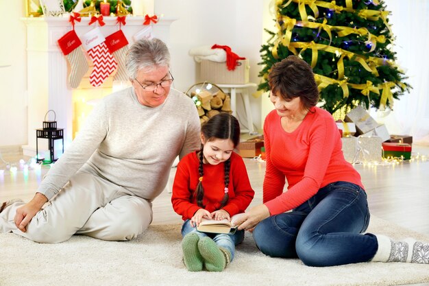 Little girl and her grandparents reading book in living room decorated for Christmas