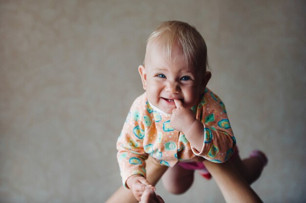 彼女の頭の上の彼女の母親の腕の中で小さな女の子は彼女の口の中で指で微笑んでいます。