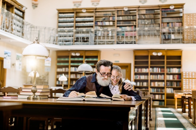 Una bambina e suo nonno barbuto senior stanno leggendo libri, seduti al tavolo con molti libri e lampada da scrivania vintage nella vecchia biblioteca antica