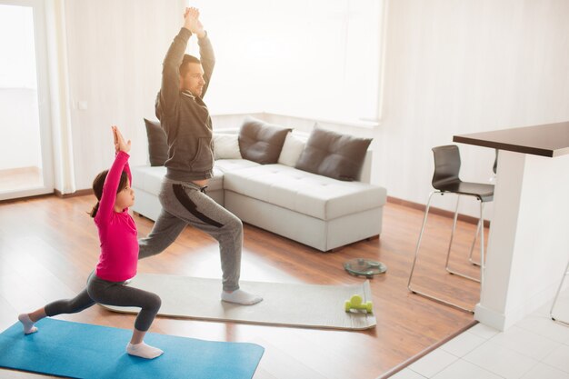 Little girl and her young father are doing lunge forward workout at home. Cute kid is training indoor. Little dark-haired female model in sportswear has exercises in room