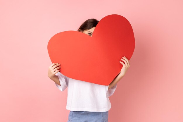 Little girl hiding face behind big red heart and looking at camera with curious prying eyes