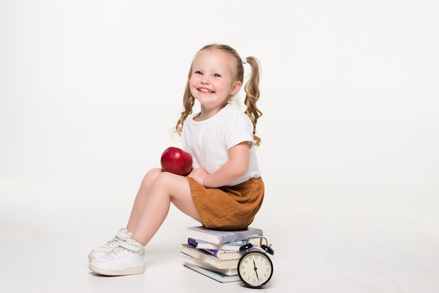 Little girl holding apple sitting on pile of books isolated