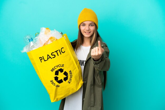 Little girl holding a bag full of plastic bottles to recycle over isolated blue background doing coming gesture