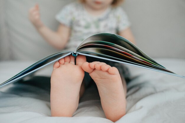 Photo little girl holding bare feet close up to camera and reading book  blurred face on background