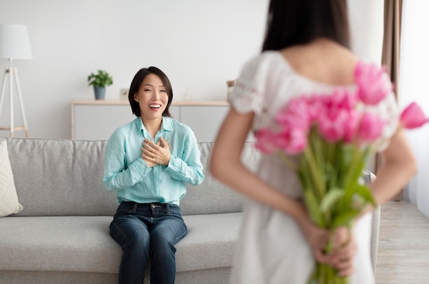 Little girl holding bouquet of tulips for happy asian granny behind her back making holiday surprise