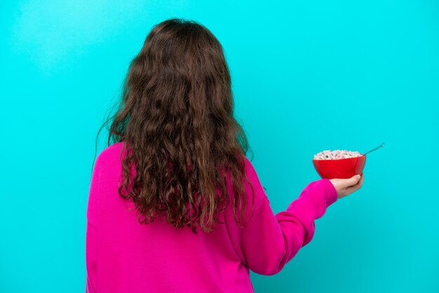 Little girl holding a bowl of cereals isolated on blue background in back position