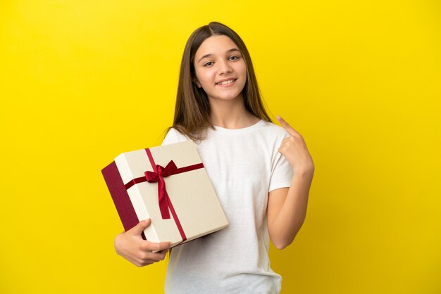 Little girl holding a gift over isolated yellow background giving a thumbs up gesture