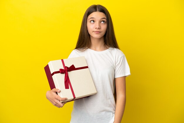 Little girl holding a gift over isolated yellow background and looking up