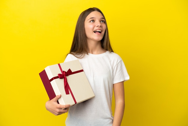 Little girl holding a gift over isolated yellow surface laughing