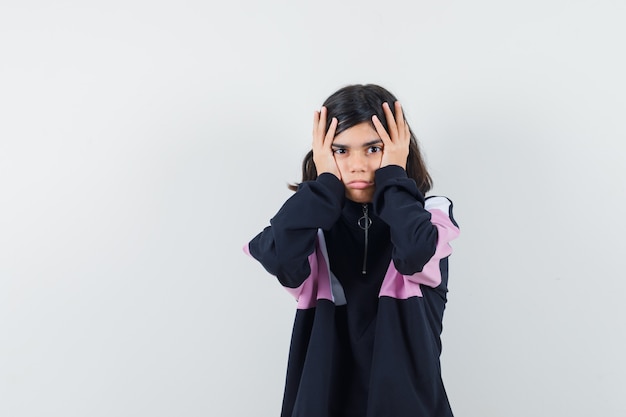 Little girl holding hands on head in shirt and looking wistful. front view.