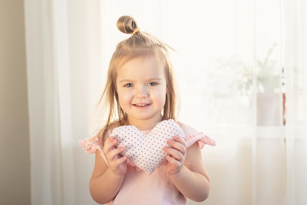 Little girl holding heart. Small heart shape pillow holding by little Caucasian girl smiling with innocence, at home. Concept of child, love and happiness, kindness, gift, handmade Valentine, familly