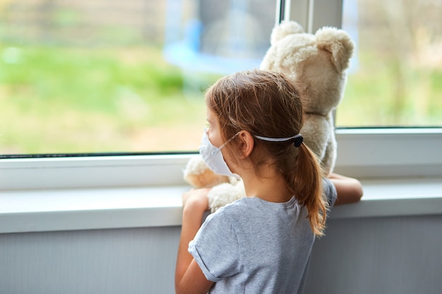 Little girl holding and hugging teddy bear in mask near the window.