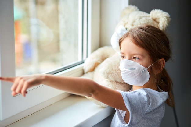 Little girl holding and hugging teddy bear in mask near the window.