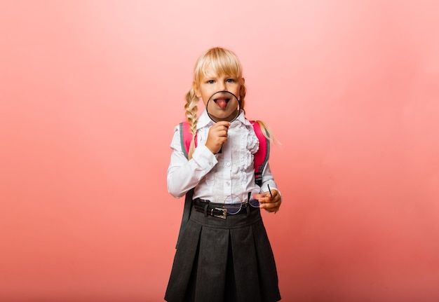 Little girl holding a magnifying glass near her mouth and showing her tongue on a pink background