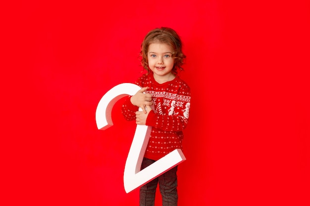 Little girl holding the number two in winter clothes on a red background, space for text