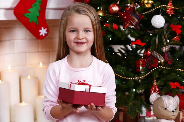 Little girl holding present box near Christmas tree on fireplace with candles background