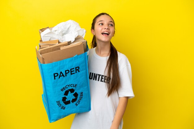 Little girl holding a recycling bag full of paper to recycle over isolated yellow background laughing