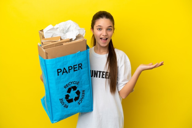 Little girl holding a recycling bag full of paper to recycle over isolated yellow background with shocked facial expression