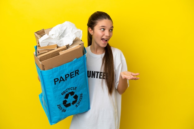 Little girl holding a recycling bag full of paper to recycle over isolated yellow background with surprise expression while looking side