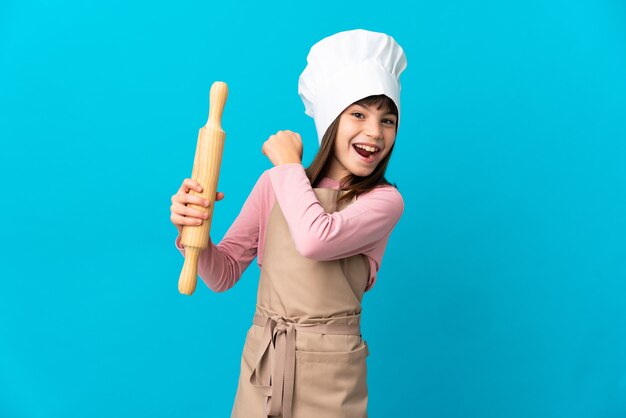 Little girl holding a rolling pin isolated on blue background celebrating a victory