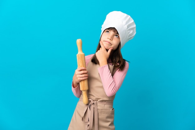 Little girl holding a rolling pin isolated on blue background having doubts