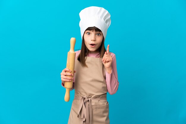 Little girl holding a rolling pin isolated on blue background intending to realizes the solution while lifting a finger up