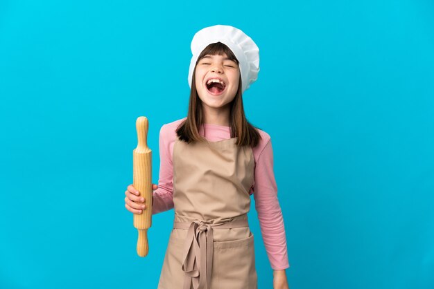 Little girl holding a rolling pin isolated on blue background laughing