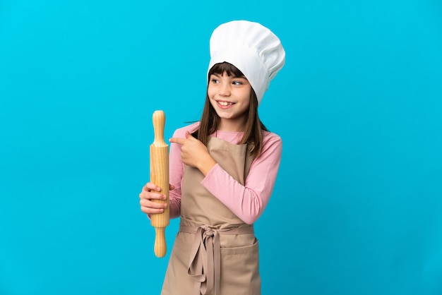 Little girl holding a rolling pin isolated on blue wall intending to realizes the solution while lifting a finger up