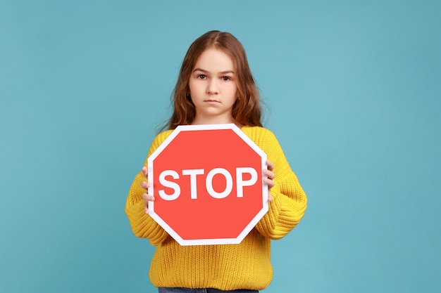 Little girl holding Stop symbol showing red traffic sign warning you about road safety rules wearing yellow casual style sweater Indoor studio shot isolated on blue background